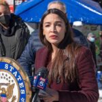Woman speaking at outdoor press event with microphones and seal