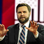 Man in suit speaking on stage with hands raised