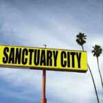 Yellow roadside sign reading Sanctuary City with palm trees