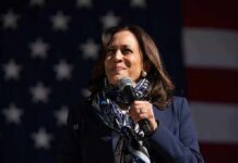 Harris’ Quiet Move, Massive Stakes Kamala Harris speaking into a microphone in front of a U.S. flag backdrop