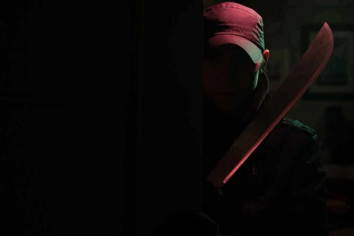 Person in cap holding a large machete in dark doorway