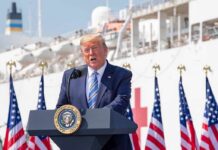 Donald Trump speaking at a podium with American flags and a ship in the background