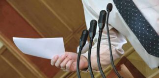 Town Hall EXPLODES – Mayor Dragged Out Speaker at podium holding paper near multiple microphones