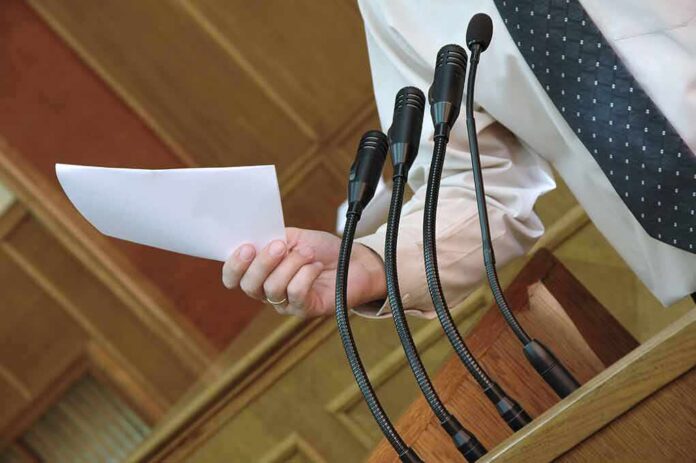 shutterstock_54523087 Speaker at podium holding paper near multiple microphones